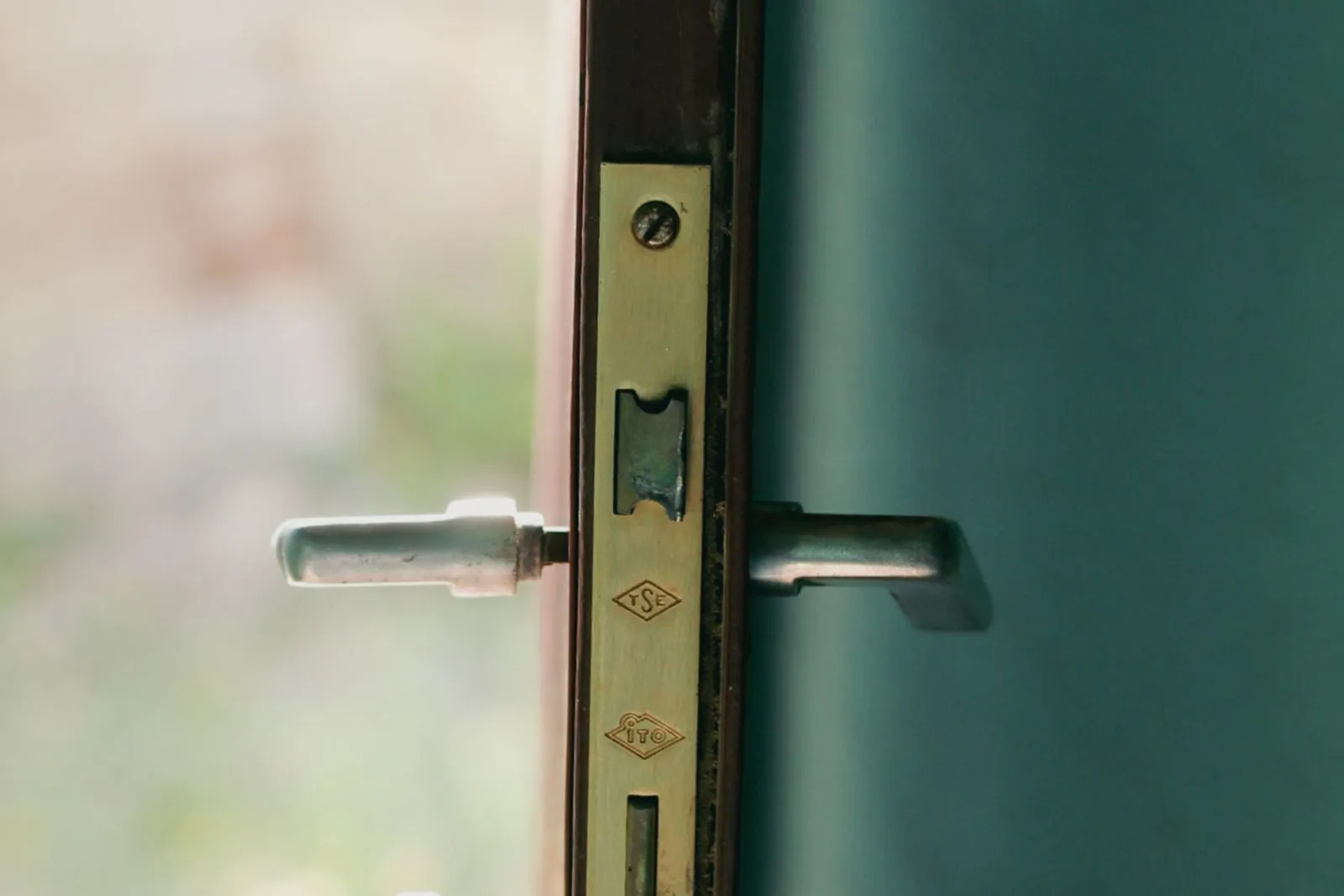 Serrure encastrée sur une porte vert sapin : têtière en laiton estampillée, clé engagée dans le canon et béquille en aluminium.